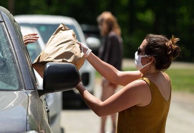 A woman wearing a mask hands a paper bag to a person in a car during a drive-through event.