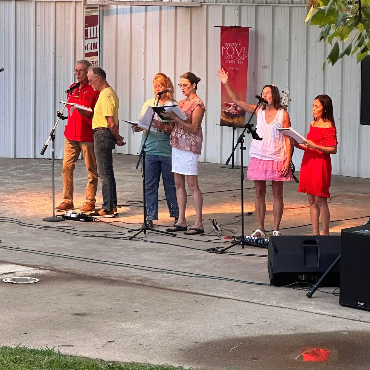 Group of six people singing on stage with microphones and music sheets.
