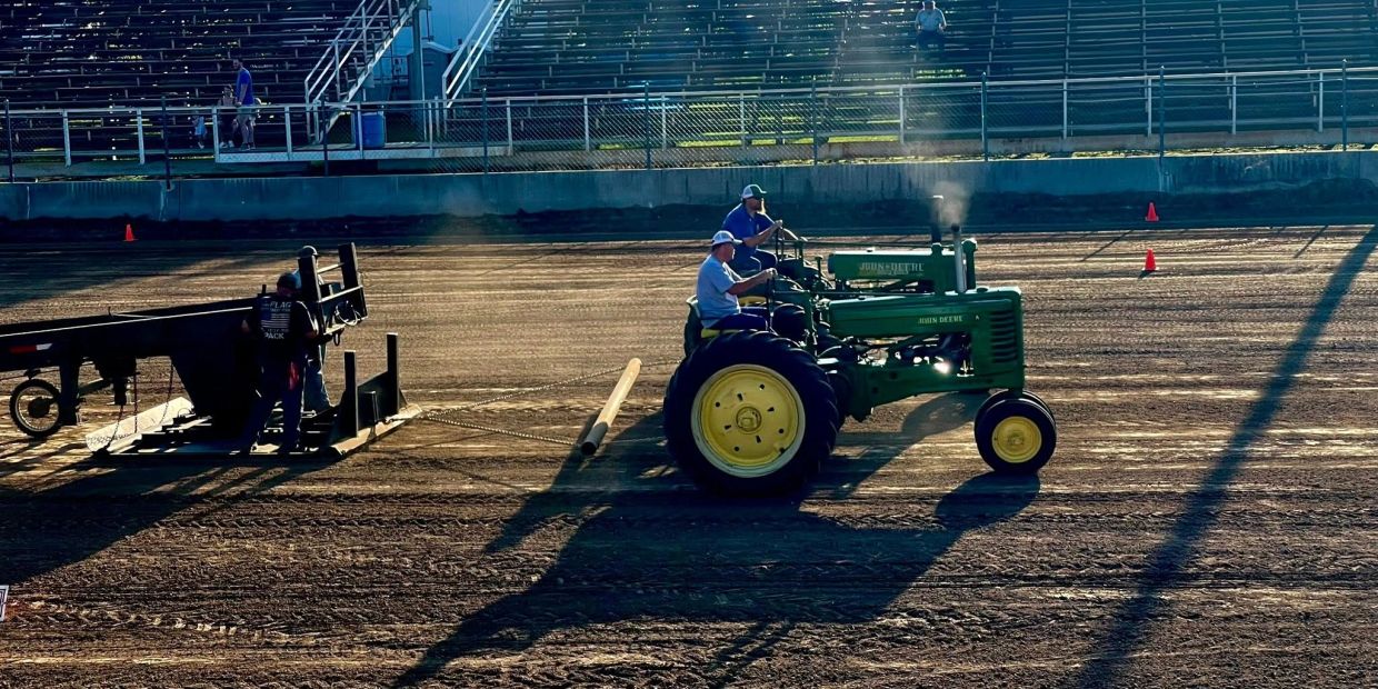 Two men driving vintage John Deere tractors on a dirt track during a sunny day.