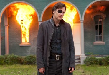 Man with eye patch stands in front of burning building.