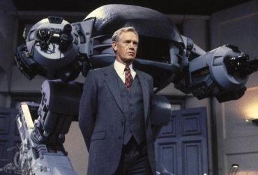 Man in a suit stands in front of a large futuristic robot.