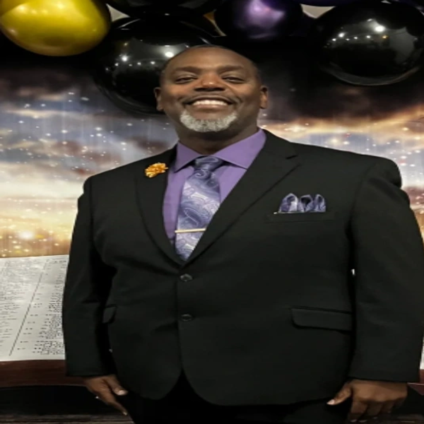 A man in a black suit smiling with a festive balloon backdrop.