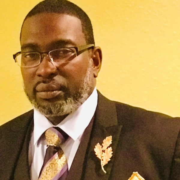 Man in a suit with glasses and a leaf pin on his lapel.