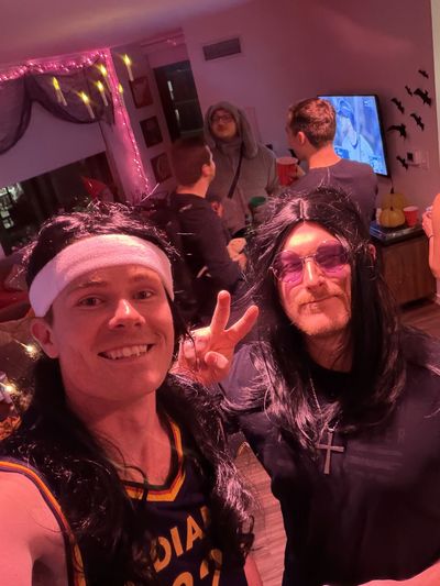 Two men in wigs pose at a lively Halloween party with decorations and drinks.
