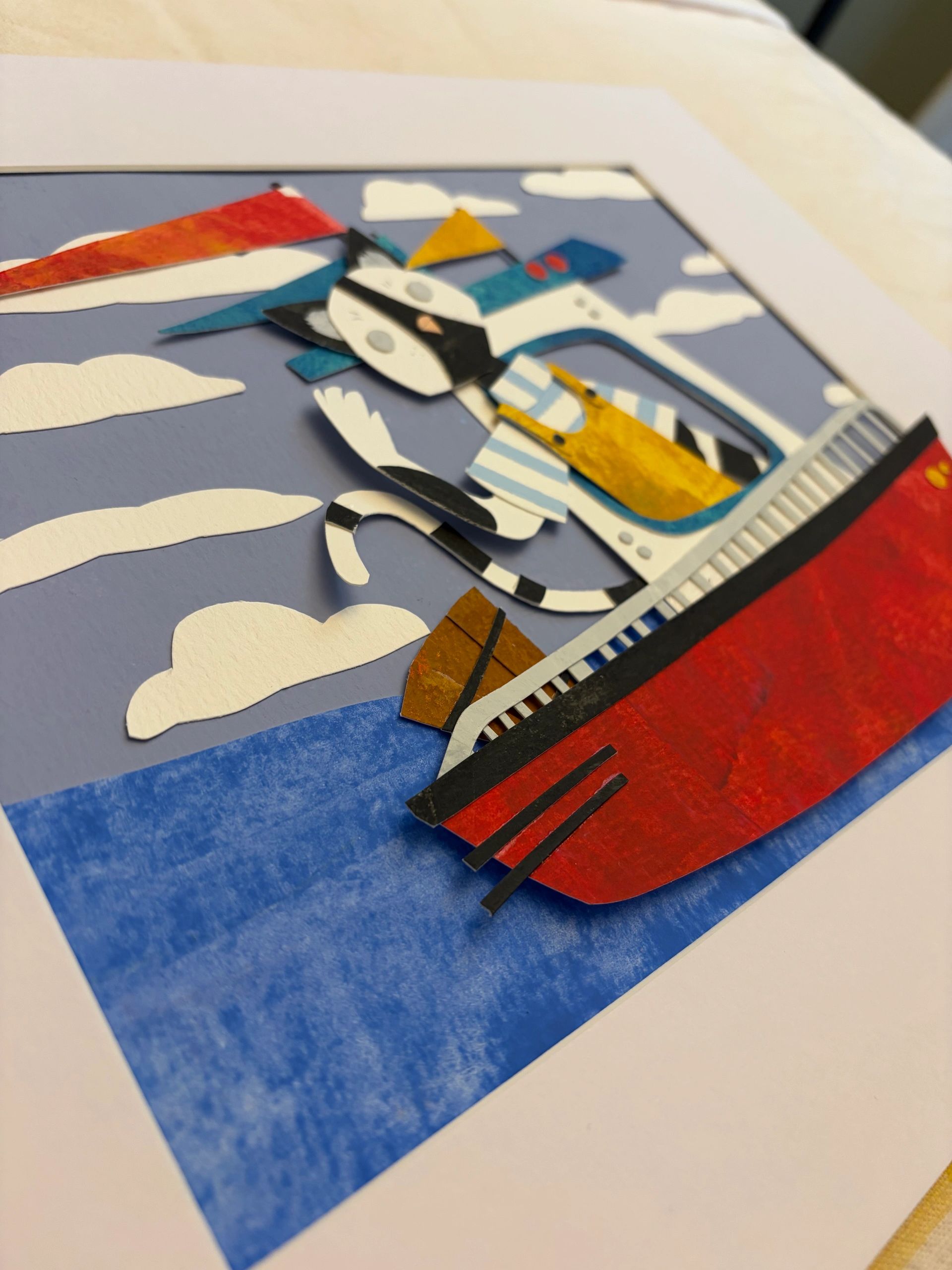 Paper cutout art of a cat on a red boat with a blue sea background.