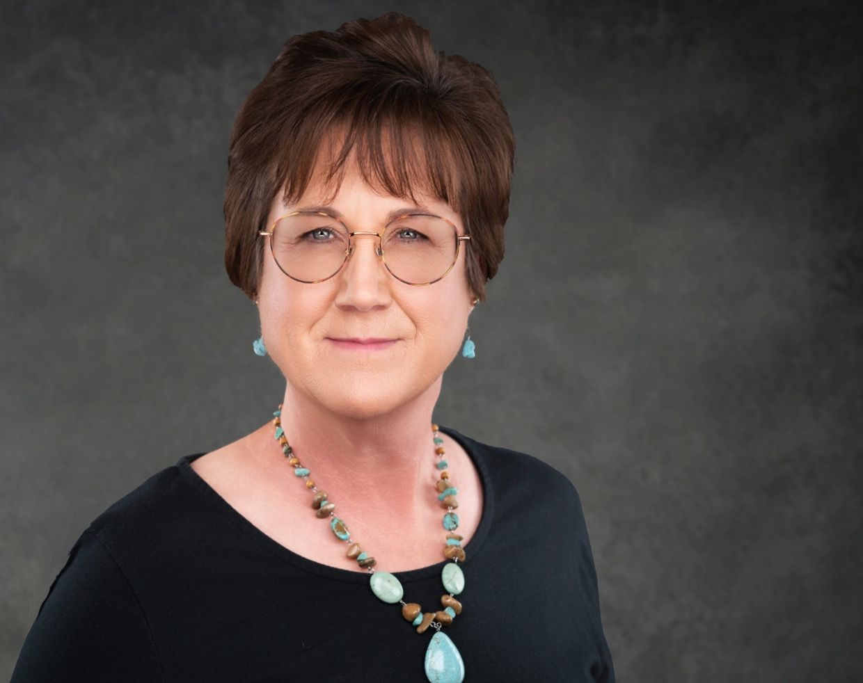 Mature woman with glasses and turquoise jewelry against a gray background.