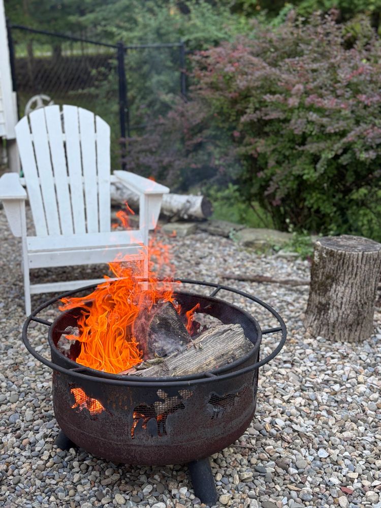 A roaring fire in a metal fire pit with animal cutouts on a gravel patio beside a white Adirondack chair.