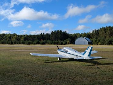 Small airplane on grass airstrip with trees and hangar in background.