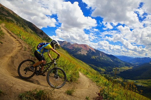 Mountain biker descends a scenic trail with mountains and clouds in the background.