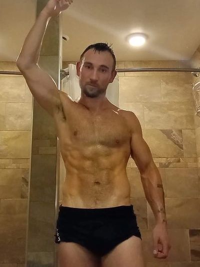 Muscular man in black swim trunks posing in a tiled shower.