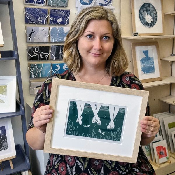 A woman in a shop holding up a Lino print of three ankles dangling in the water.