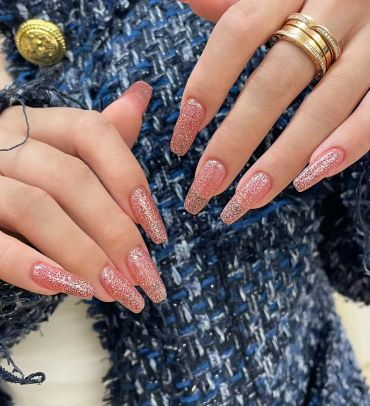 Hands with long, glittery pink nails and gold rings on a textured blue fabric background.