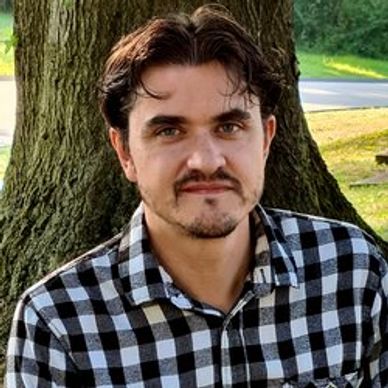 Man in a black and white checkered shirt sitting outdoors by a tree.