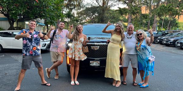 Group of six people happily posing with shaka signs in front of a black Cadillac in a tropical setting.