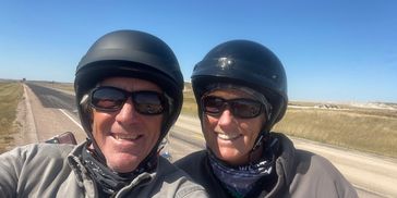 Two people wearing helmets and sunglasses smiling on a motorcycle ride.