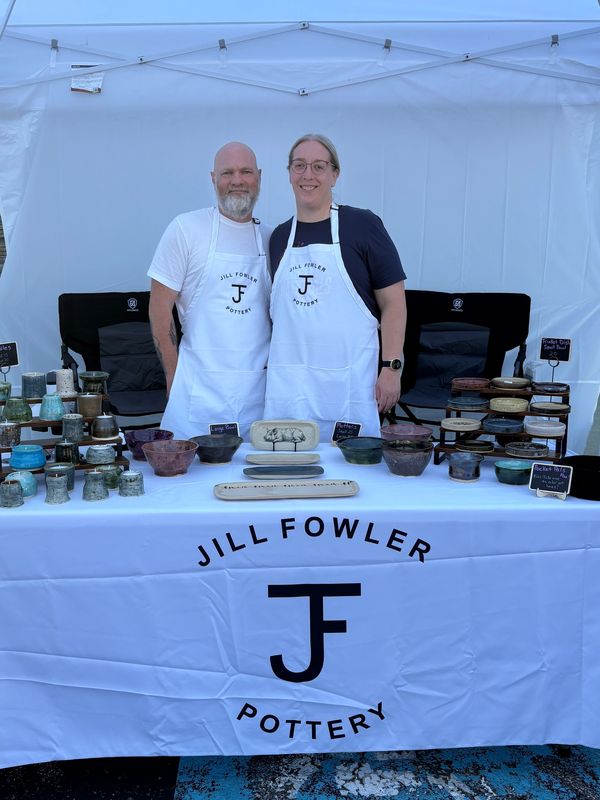 Two people with aprons stand behind a pottery display table at a market stall.