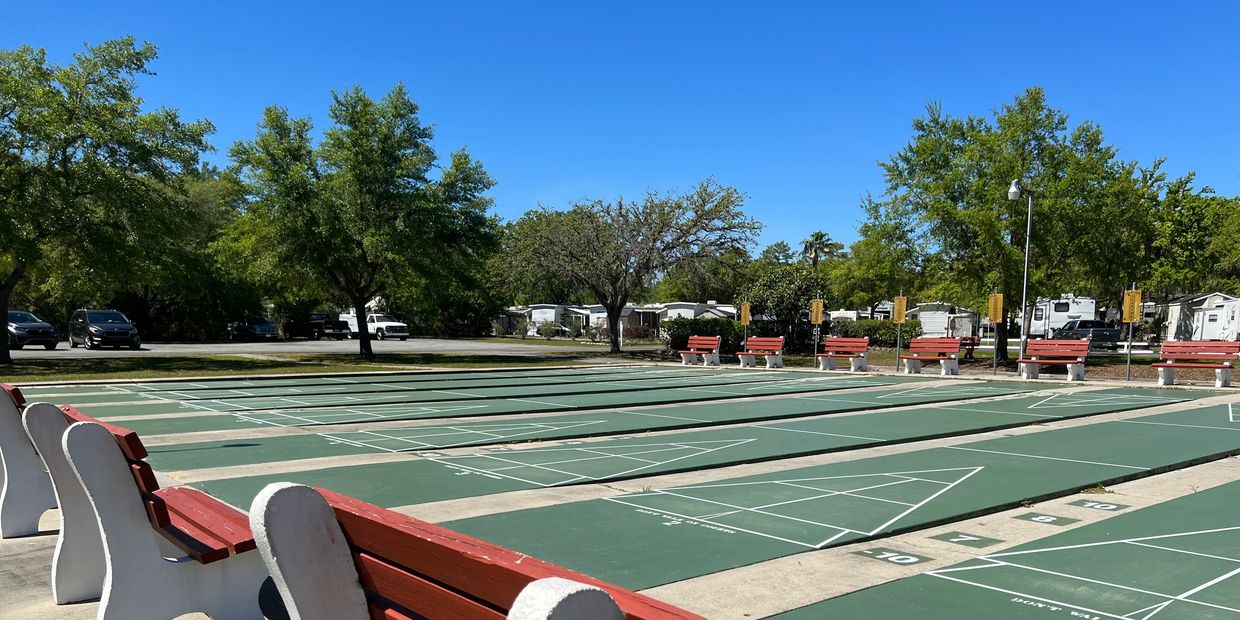 A row of outdoor green shuffleboard courts lined with red and white park benches, surrounded by tree
