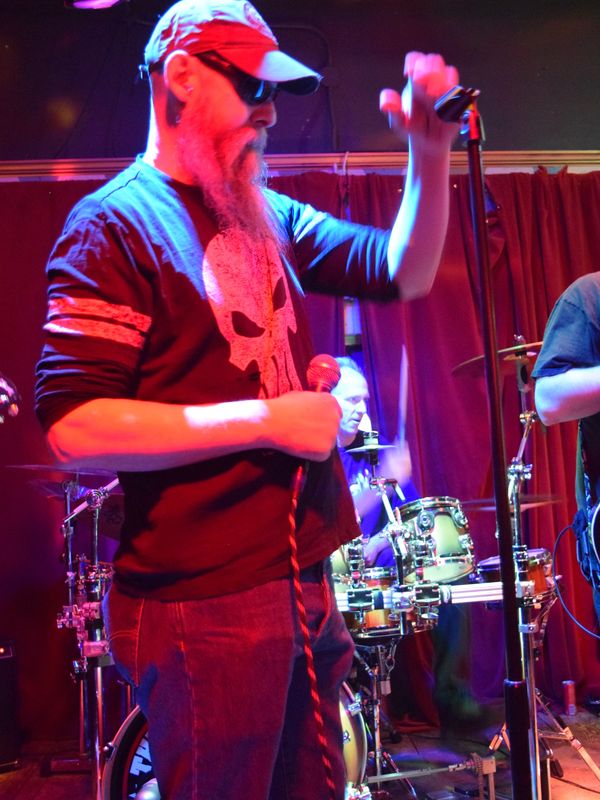 Bearded man in sunglasses passionately singing on stage with a band.