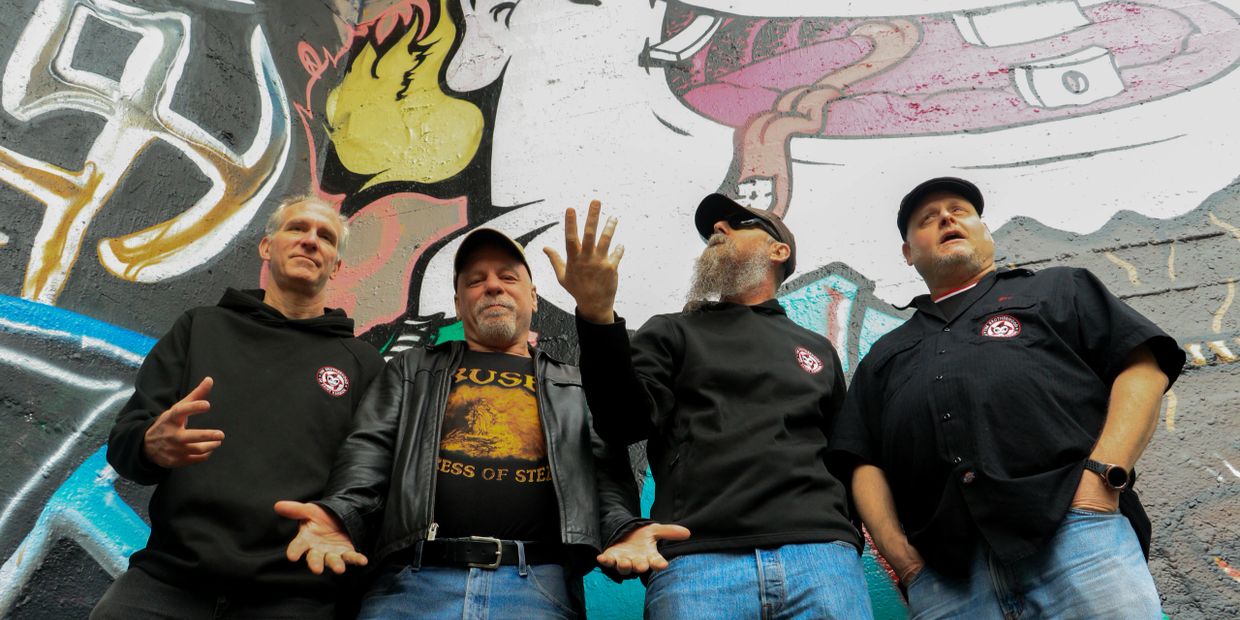 Four men posing in front of colorful graffiti, some wearing black hoodies and hats.