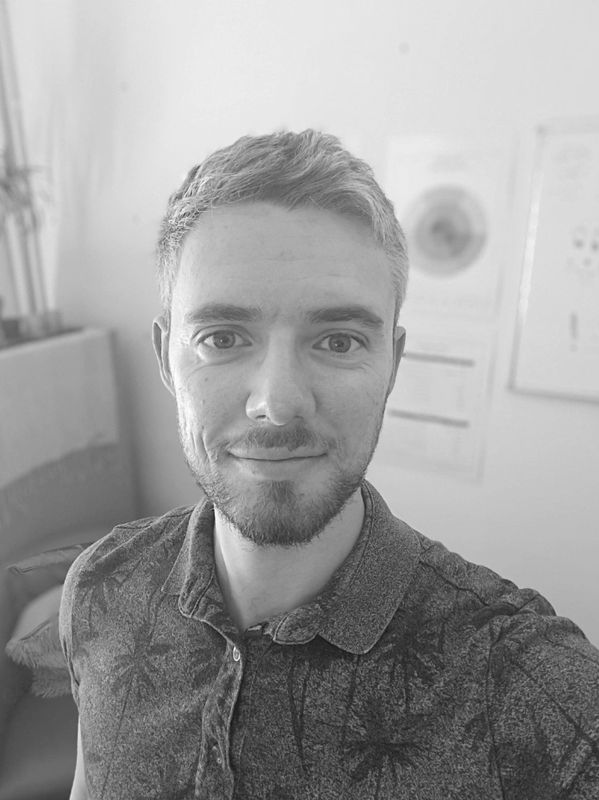Smiling young man with short hair and beard in a casual shirt taking a selfie indoors.