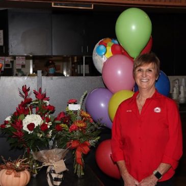 Smiling woman in red shirt with colorful balloons and flower bouquets.
