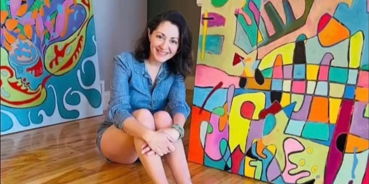 Young woman sitting on floor smiling beside colorful abstract paintings.