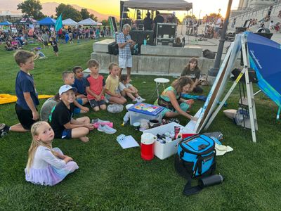 Children sitting on grass watching a live painting demonstration outdoors.