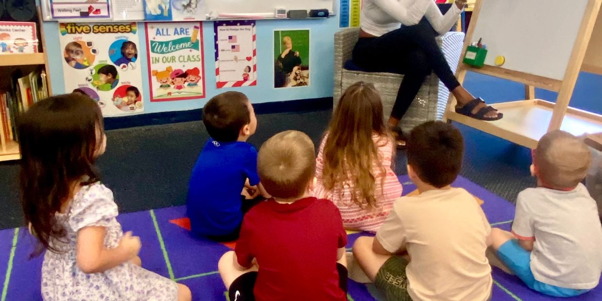 Children sitting on a classroom carpet listening to a teacher.