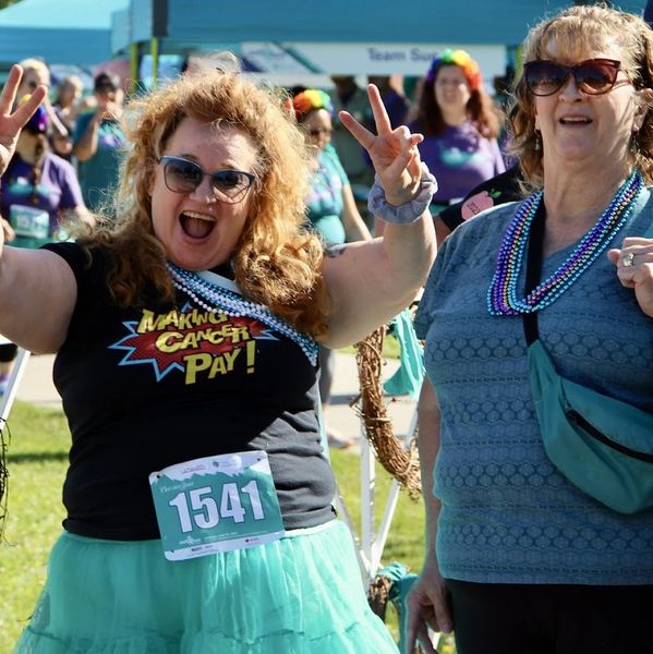 Two women at a cancer awareness event, one joyfully posing with peace signs.