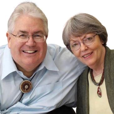 Smiling elderly couple wearing glasses and necklaces, posing closely together.