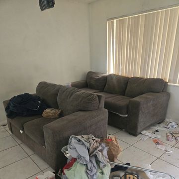 A messy living room with two brown sofas and scattered clothes and trash.