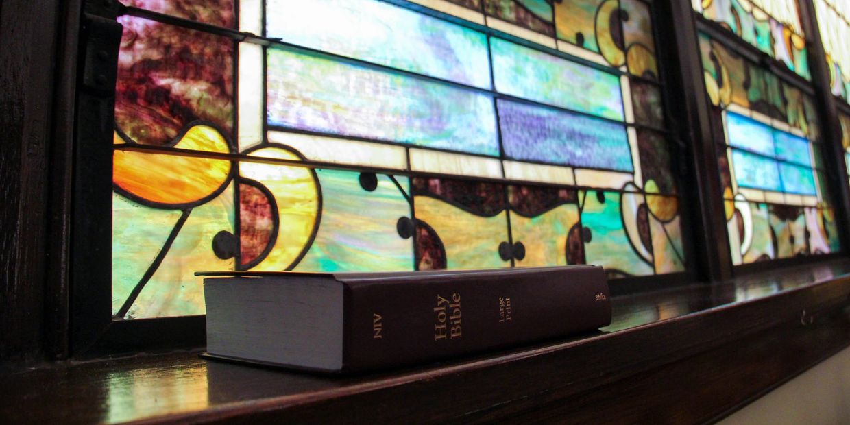 A Holy Bible rests on a wooden ledge in front of a colorful stained glass window.