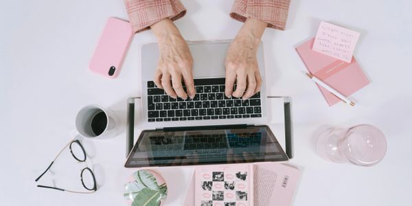 Person typing on a laptop surrounded by pink-themed stationery and coffee.