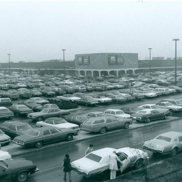 Vintage parking lot filled with classic cars near a large shopping center.