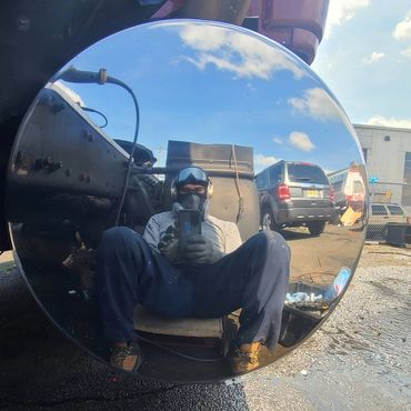 Person reflected in a large round mirror outdoors, wearing a mask and work clothes.