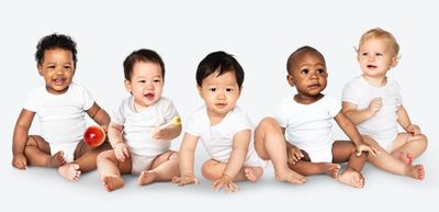 Five diverse babies sitting in white onesies against a plain background.