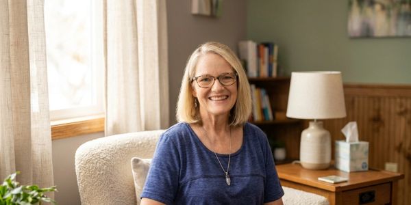 Smiling woman with glasses sitting comfortably in a cozy, well-lit room.
