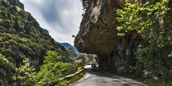 Motorcyclist rides through a narrow mountain road with rocky cliffs and greenery.