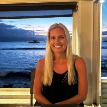 Smiling woman with long blonde hair sitting by the sea at sunset.