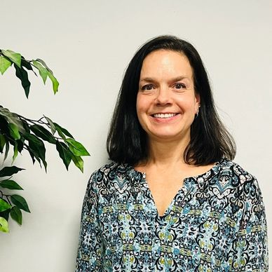 Smiling woman with dark hair in patterned blouse by a plant.