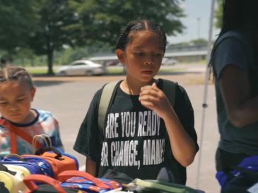 Child looks at backpacks on a table outdoors.