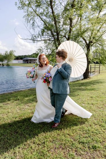 Couple in wedding attire walking by a lakeside with an umbrella.