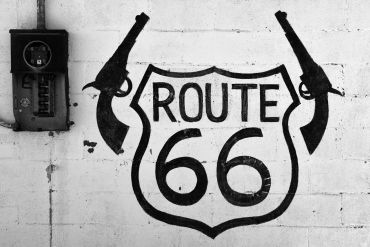 Black and white Route 66 sign with guns on a wall.