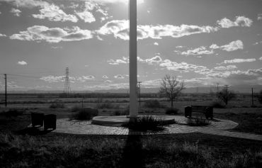 Black and white image of a flagpole and benches in a rural setting under a cloudy sky.