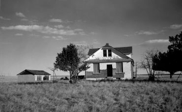 Black and white photo of an old abandoned house and a small shed in a grassy field.