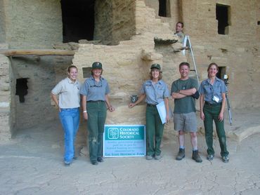 Five people stand in front of an ancient stone structure with a Colorado Historical Society sign.