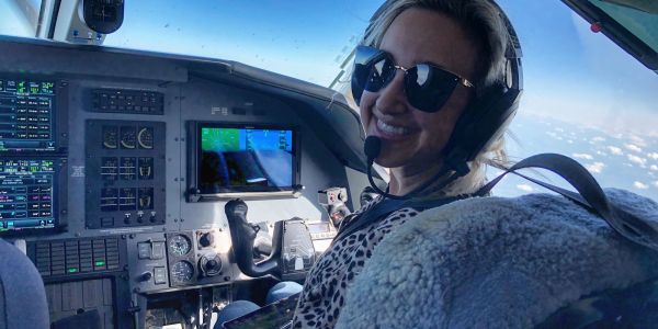 Smiling woman pilot wearing headset and sunglasses inside airplane cockpit.