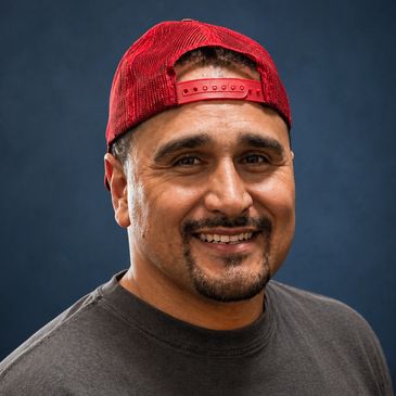 Smiling man wearing a red backward cap and dark shirt against a blue background.