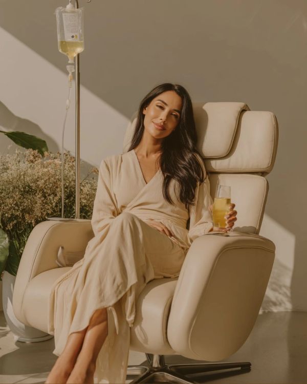 Woman in beige robe relaxing in a cream chair with a drink and IV drip.