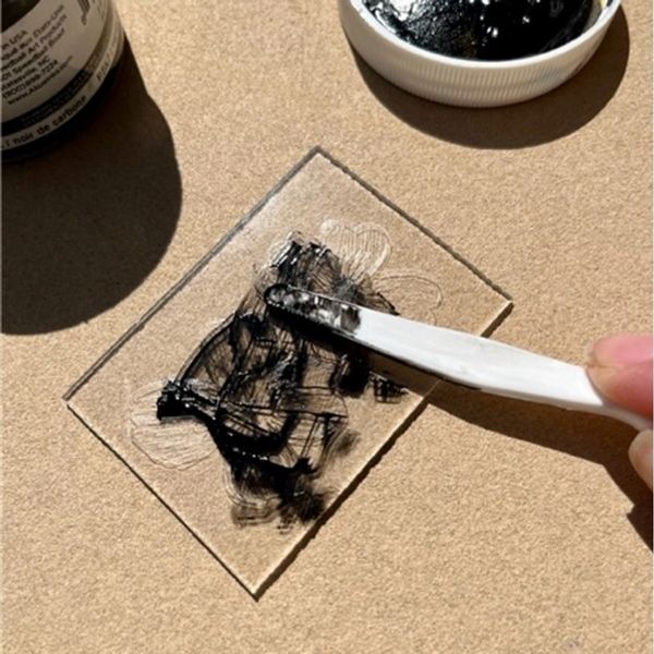 A hand holding a palette knife spreading ink into an itaglio printing plate on a brown background